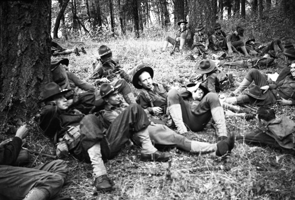 About a dozen soldiers sitting on the ground in a heavily wooded area.  Most of them are sitting up against trees and in full military gear.