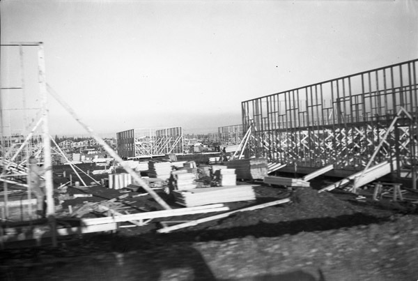 Multiple buildings in the very early stages of construction.  Only one wall per building has been raised, and large wood piles are around each of them.
