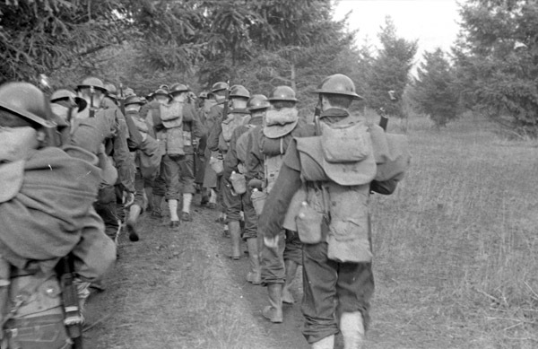 A group of a dozen soldiers marching away from the perspective of the camera. They are all in full military gear with backpacks and sleeping bags.
