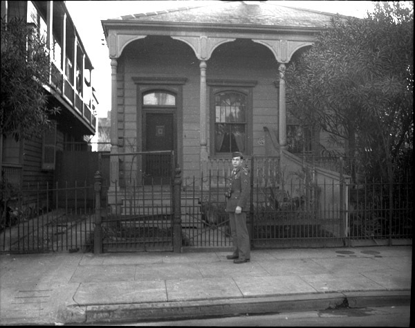 A soldier in dress uniform and a camera hanging from their neck standing in front of a single-level house.