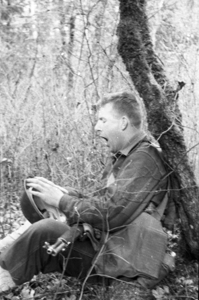 A soldier sitting on the ground with their back against a tree.  They look to be yawning. and holding their helmet in their lap.