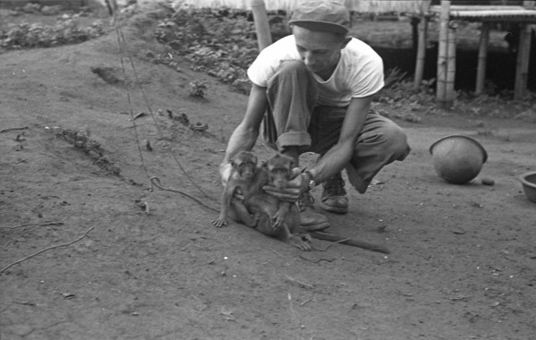 A monkey, named Jocko, sitting on the ground with another monkey beside it.  A man in short sleeves holds both of them so they face the camera.