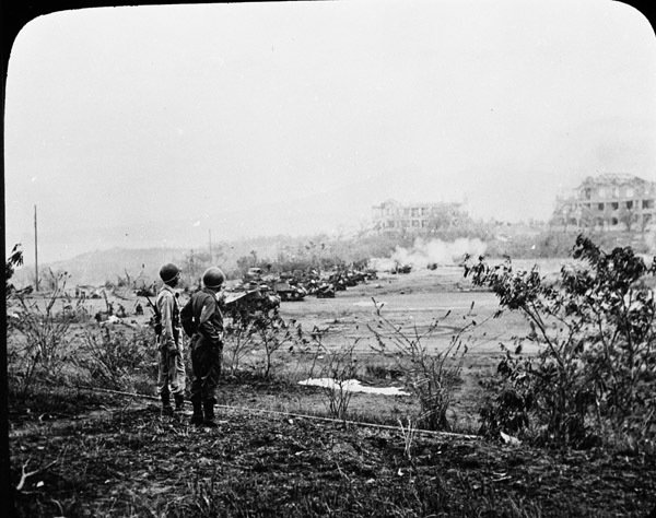 Two military officers look over a field of artillery.  Smoke and destroyed buildings are in the background, likely from a recent battle.