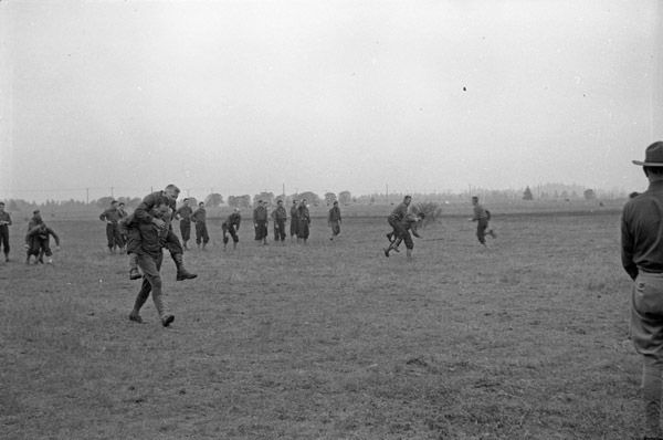 A small group of soldiers in a field giving other men piggy-back rides and smiling. They appear to be racing, while other men look on the race.