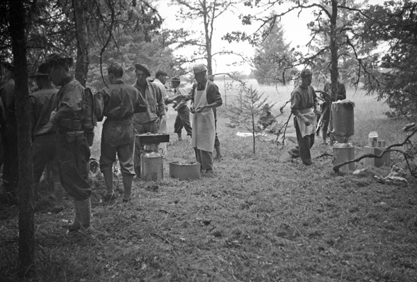 A group of soldiers in a wooded clearing standing around pots of food. The man in the center is wearing a white cook's apron and hat.