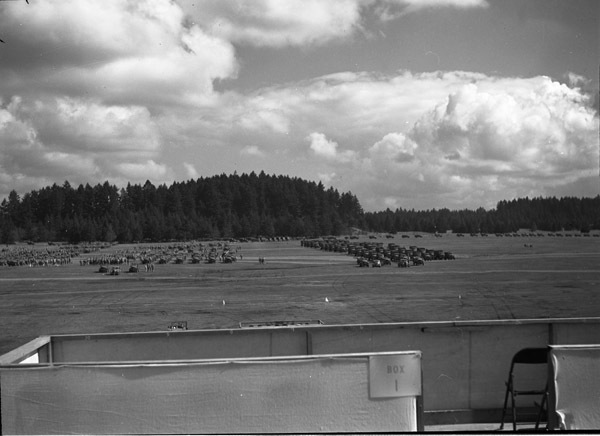 An elevated landscape view of military trucks and soldiers in lines in a wooded clearing. A seating area with a sign labeled "Box 1" and containing a folding chair is in the foreground, facing the soldiers and vehicles.