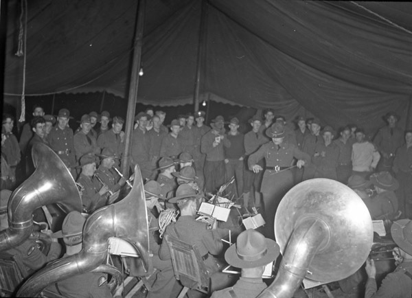 A military band plays inside a large tent.  One man is conducting, and the entire background is filled with more soldiers watching the performance.
