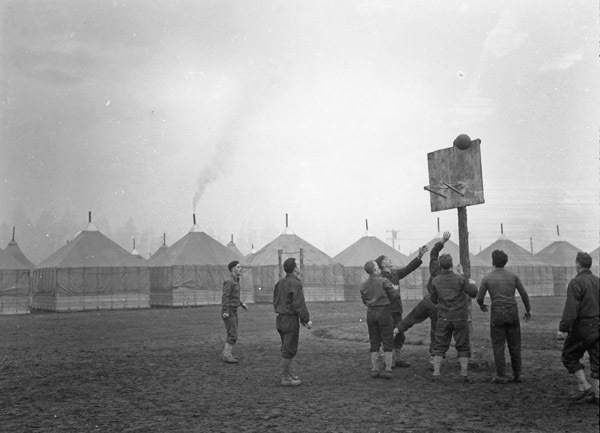 Eight soldiers playing basketball around a hoop. The ball can be seen in mid-air hitting the backboard. Small houses or tents line the background.