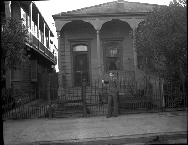 A soldier in dress uniform with their arm leaning on a fence in front of a single-level house.