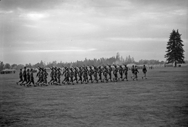 A couple dozen soldiers marching in formation with rifles resting on their shoulders.  A tent camp can be seen in the background.