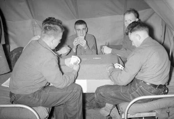 A group of five soldiers sitting on a pair of cots and playing card games on a card table that is set up between them.