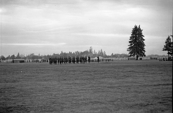 A distant view of a dozens or more soldiers in formation while officers walk in front of them. A tent camp and milling soldiers can be seen in the background.