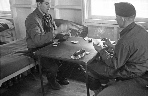 Two soldiers sitting on bunks with a card table between them, playing cards and smoking.
