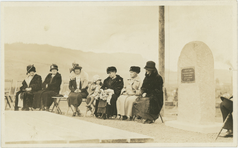 A group of six women and one young girl sitting on chairs next to a dedication plaque.