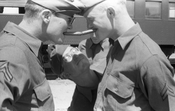 Two soldiers humorously face each other while faux-scowling.  Each of them has a cigar in their mouths, and the cigar ends are touching each other.  Another soldier is behind them and peering at the seen amusedly.