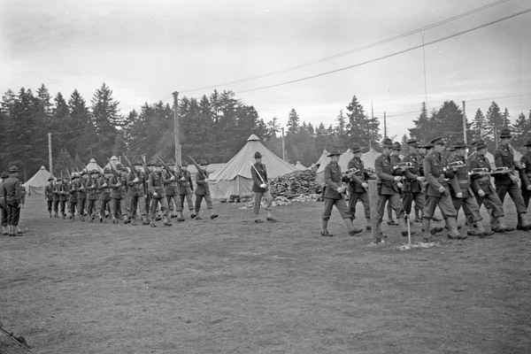 Two large groups of soldiers marching in formation next to a tent camp. The far right group is in front and holding musical instruments. The leftmost group in the back has rifles resting on their shoulders.