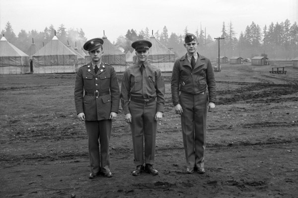 Three soldiers in military dress uniform standing shoulder-to-shoulder posing for the camera. Tents and fir trees can be seen in the background.