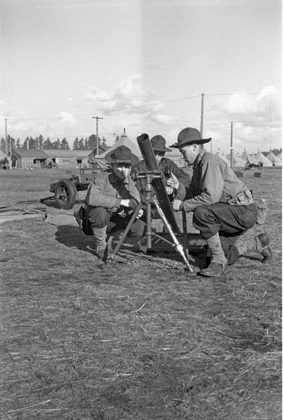 Three soldiers, each squatting down on one knee, huddled around a mortar.  A tented camp can be seen in the background.