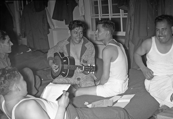 A group of five Hispanic soldiers sitting or laying on the floor in casual clothes and smiling.  The soldier in the middle is playing a guitar. 