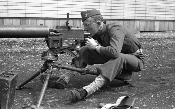 A soldier sitting on a seat behind a tripod-mounted machine gun. He is looking through the sights of the gun.