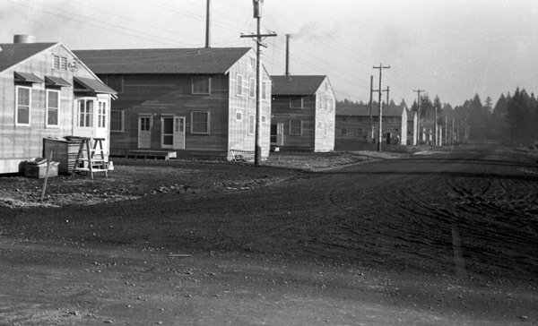 A line of two-story houses on the left, with a dirt road on the right.  Trees are in the background.