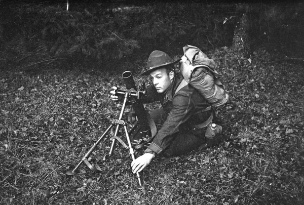 A soldier surrounded by bushes and trees squatting down next to a mortar and adjusting it on its tripod.