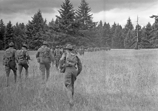 Dozens of soldiers walking in a marching line walking away from the camera through a clearing surrounded by woods.