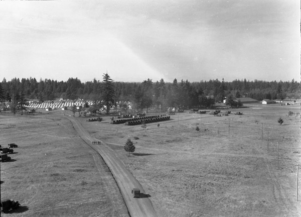 An view from on high and at a distance of a semi-permanent military camp in a wooded clearing. A dirt road is in the foreground leading into the camp.
