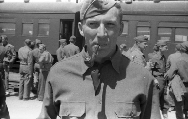 A soldier wearing a garrison cap and smoking a pipe.  Other soldiers and a pair of train cards can be seen in the background.
