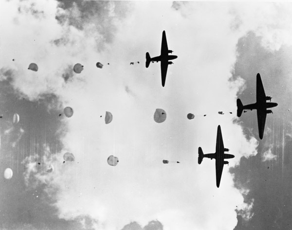 Three military planes in the sky as seen from directly below.  There are numerous paratroopers in the sky behind the planes in various stages of deploying their parachutes.