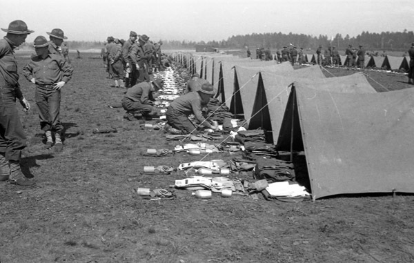 A group of soldiers kneeling or standing and facing a line of tents. In front of the tents are neatly organized groups of equipment, most likely the contents of the tent and the soldiers' belongings.