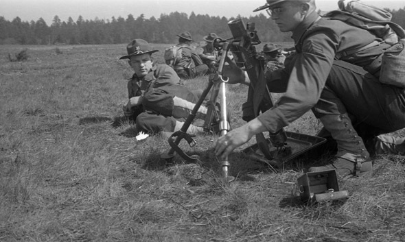 A soldier kneeling next to a mortar and adjusting its tripod. Another soldier is laying on the grass beside him watching, and another small group of soldiers can be seen nearby in the background.