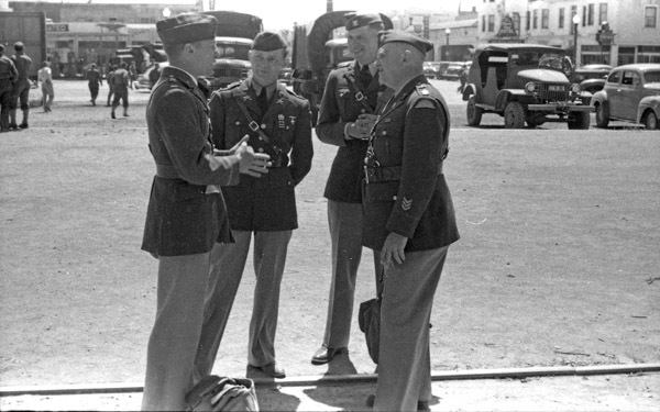 Three soldiers listen to a fourth man talking to them. All of them are in full military dress uniforms. Various military trucks and jeeps are in the background.