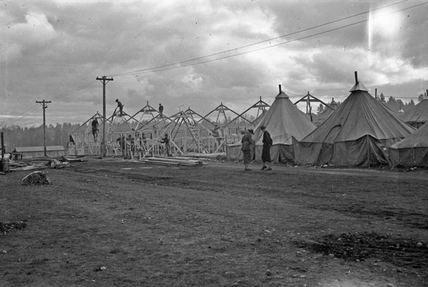 Soldiers working on skeletal wooden structures that make up large tents.  Two completed tents can be seen in the foreground.