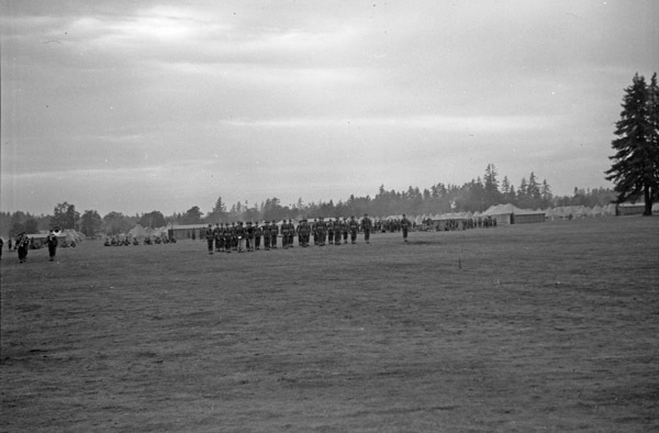 A distant view of a dozens or more soldiers in formation while officers walk in front of them. A tent camp and milling soldiers can be seen in the background.