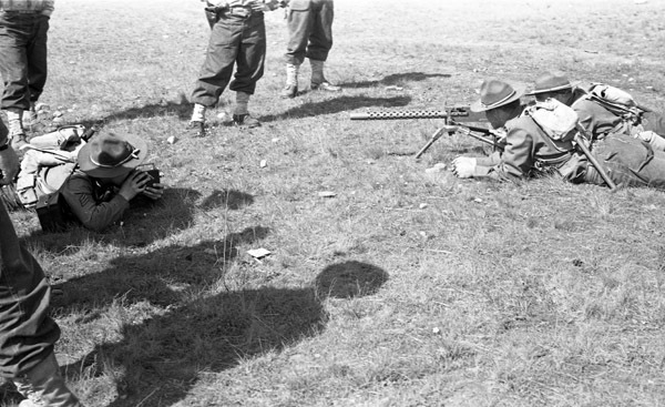 Two soldiers laying down and aiming a large machine gun that is resting on a tripod.  Opposite them is a man laying down facing them and recording them with a camera.  All three are surrounded by other soldiers, who can only be seen from the waist down.