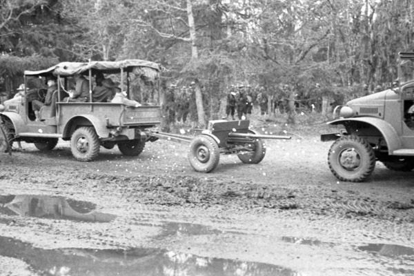 A military truck towing an artillery weapon.  Another truck is following it, and soldiers are seen against a tree line in the background.