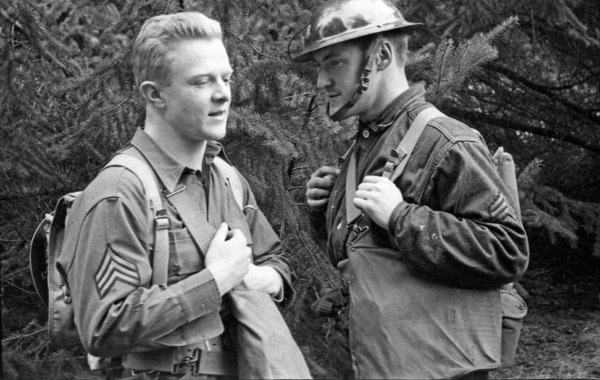 Two soldiers talking with one another.  The one on the left is not wearing their helmet, while the one on the right is wearing theirs.  Trees line the background.