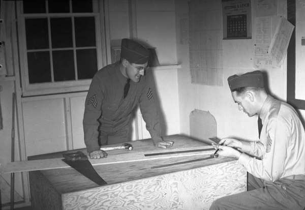 Two soldiers in dress uniform leaning over a carpentry table with various tools on it.