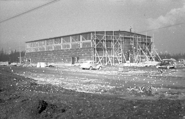 A building under mostly complete construction, with scaffolding on the nearest side. Two care are parked in the foreground.