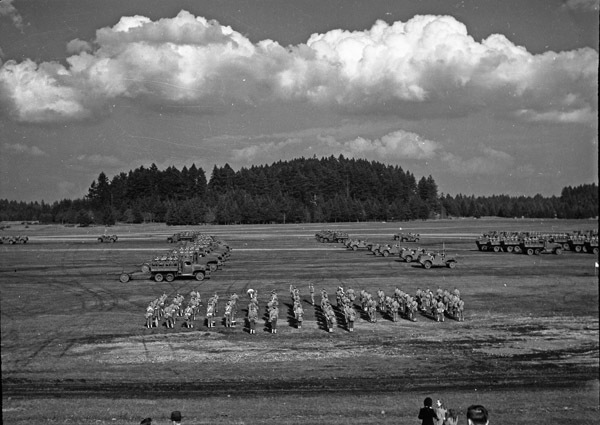 An elevated landscape view of military trucks and soldiers in lines in a wooded clearing. A few onlookers can be seen in the foreground.
