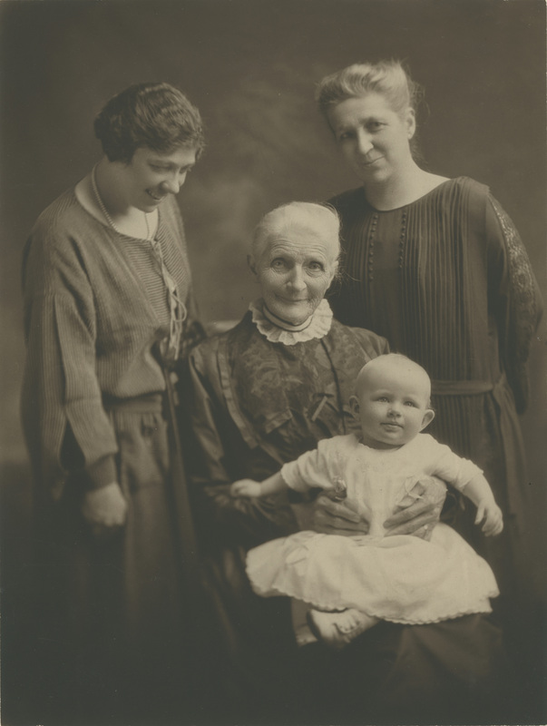 Two women standing around an older woman holding a baby girl in her lap.
