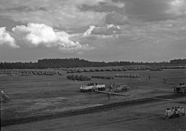 An elevated landscape view of military trucks and soldiers in lines in a wooded clearing. A civilian vehicle is in the foreground, and a stage is set up in the center.