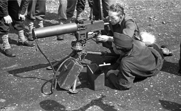 Two soldiers looking and aiming with a large, ground-mounted machine gun. One is looking from the rear, seeming to aim, while the other lays on the ground watching.