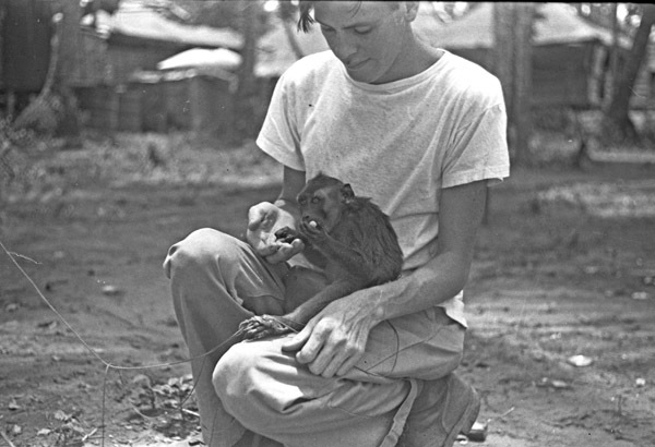 A monkey, named Jocko, sitting in the lap of a squatting young man.  The monkey is eating something being given to it by the man.