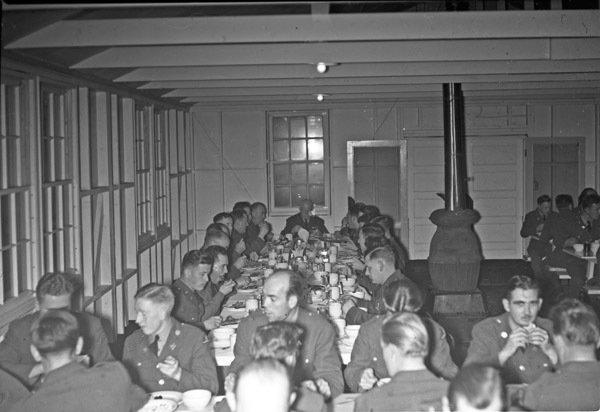 A group of soldiers sitting at a long table having a dinner.   A similar table can be seen partially in the foreground, also occupied by dining soldiers. 