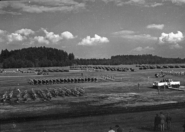 An elevated landscape view of military trucks and soldiers in lines in a wooded clearing. A stage is set up to the right