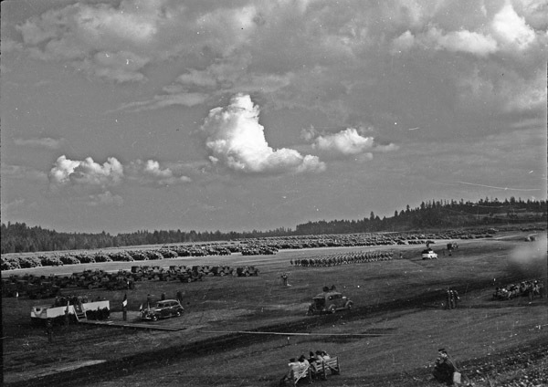An elevated landscape view of military trucks and soldiers in lines in a wooded clearing. A few civilian vehicles are in the foreground, and a stage is set up on the left.