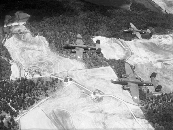 Five bombing planes flying over a moderately forested grasslands, from the view of another airplane.