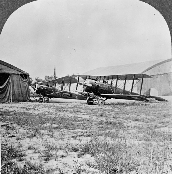 Two biplanes sitting in an airfield, with a tent to their left and an airplane hanger in the right background.